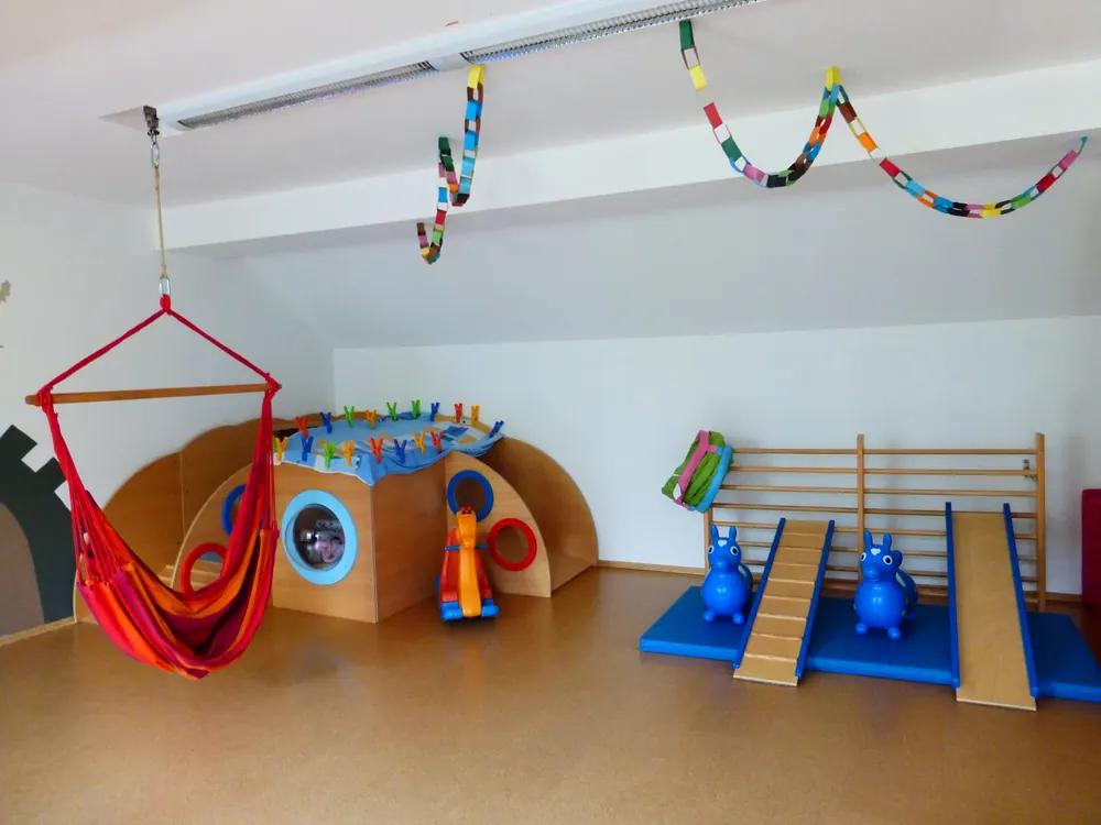Bewegungsraum mit roter Hängeschaukel, Holzspielhaus, Sprossenwand und Rutschbrettern