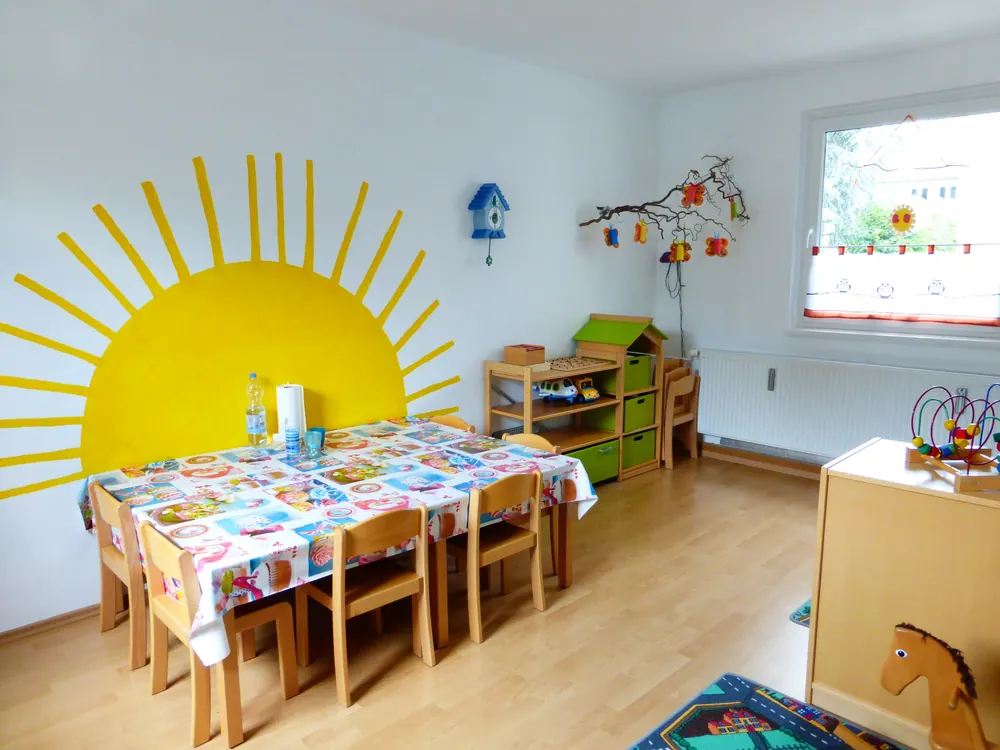 Gruppenraum mit gedecktem Kindertisch vor einer gemalten Sonne an der Wand