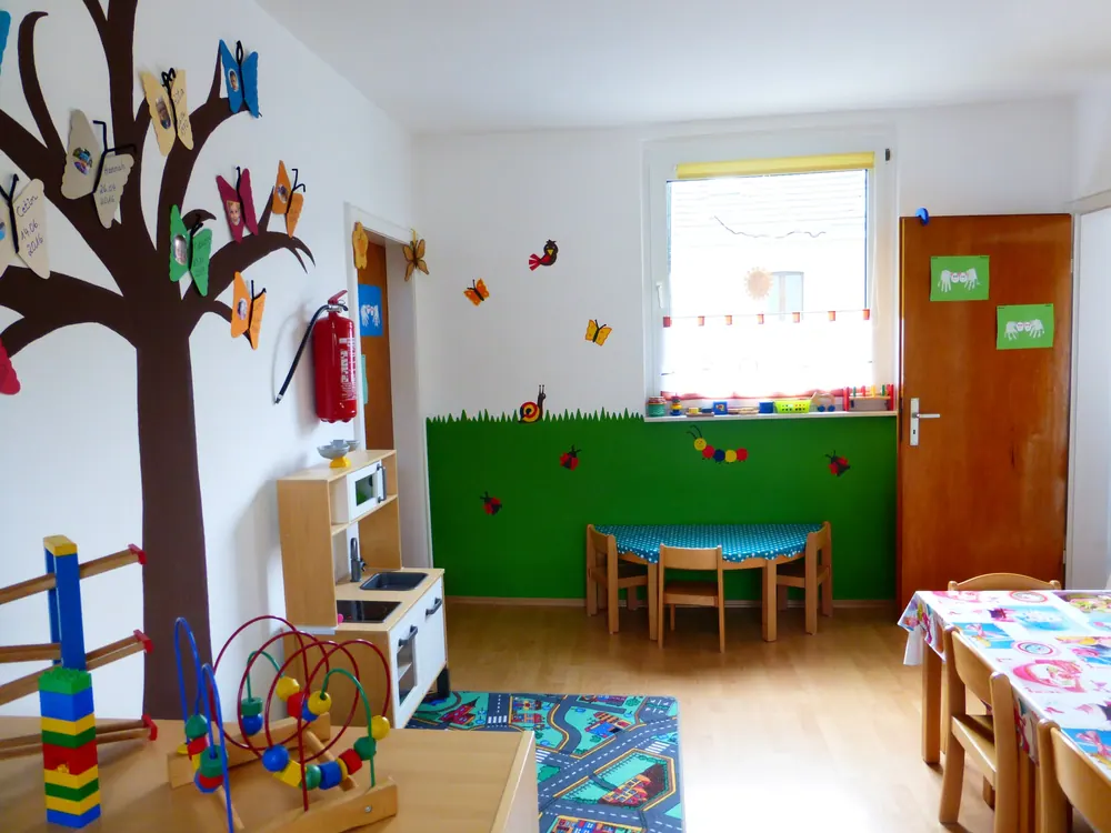 Gruppenraum mit Spielküche, kleinem Tisch und einem gemalten Baum mit Fotos der Kinder