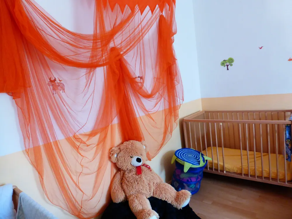 Schlafraum mit Gitterbett, großem Teddy und orangefarbenem Baldachin